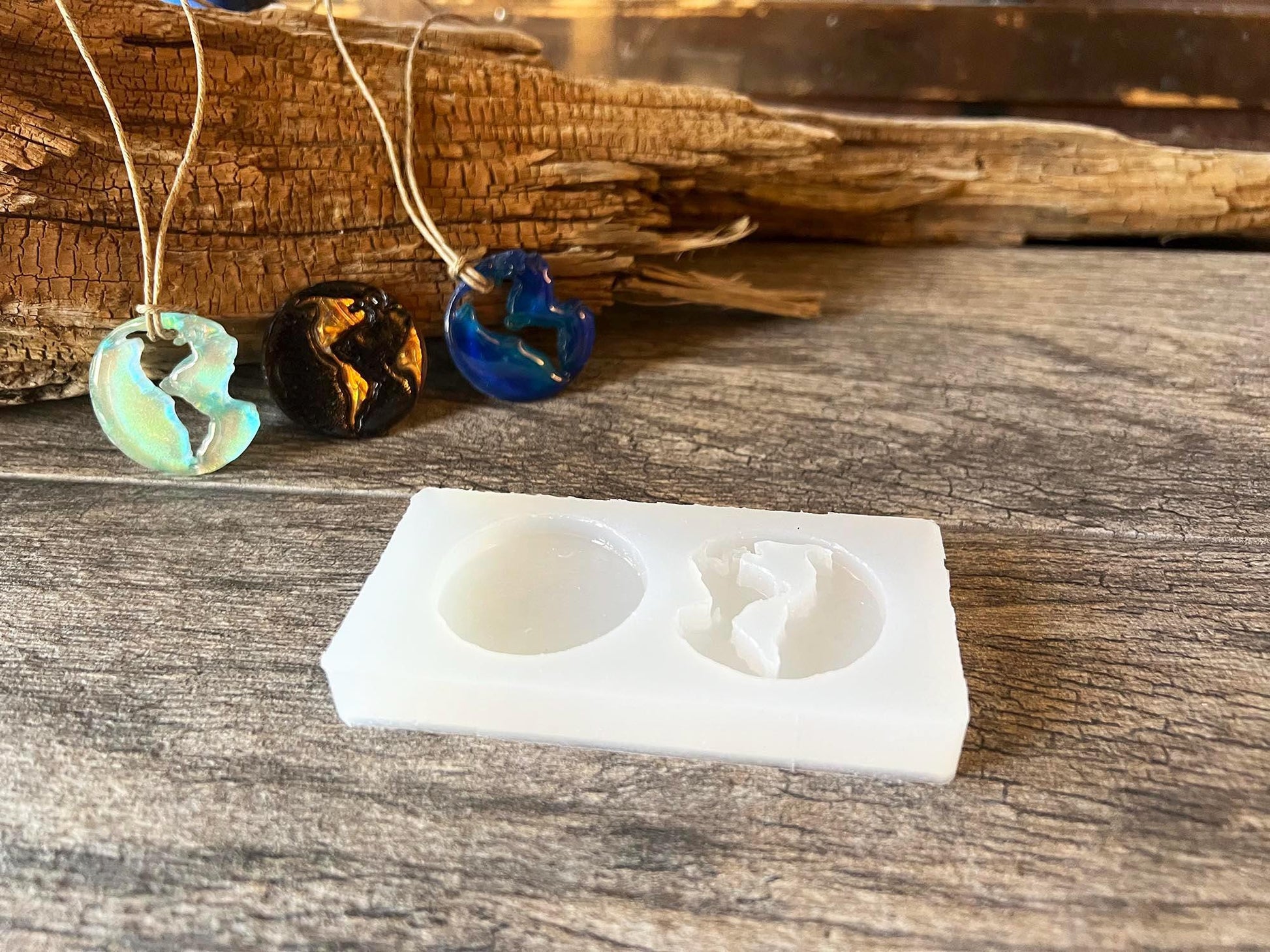Silicone mold with two round cavities displayed on a wooden surface, one solid circle and one open-continent earth design; finished resin earth pendant necklaces in blue, black, and iridescent tones hang above on natural cord.